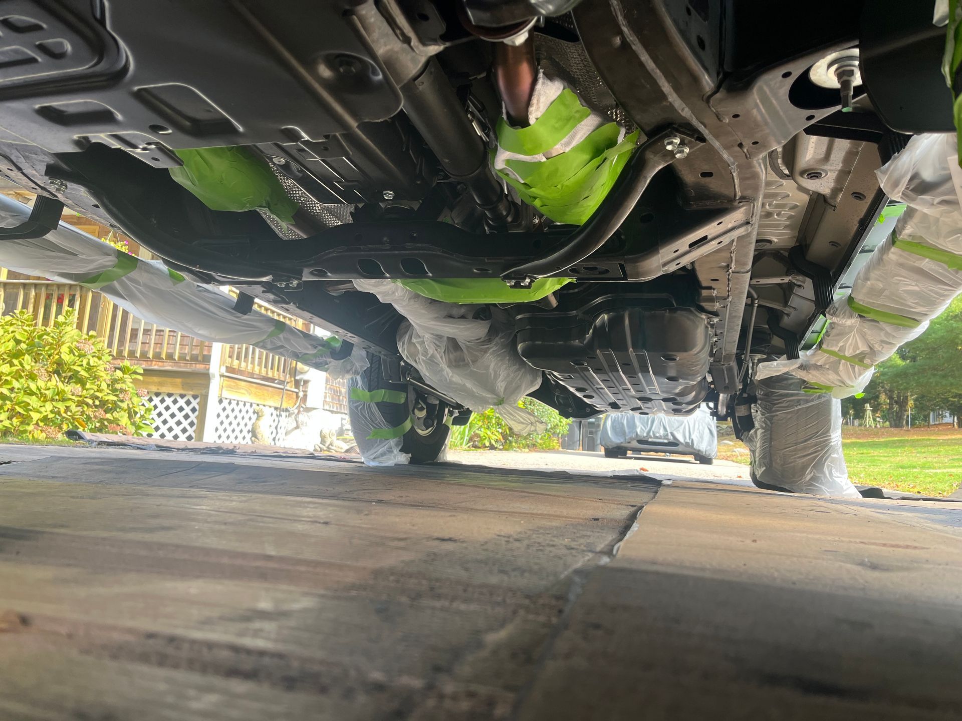 Underside of a vehicle being painted with green tape masking sections. The work is being done on a concrete surface.
