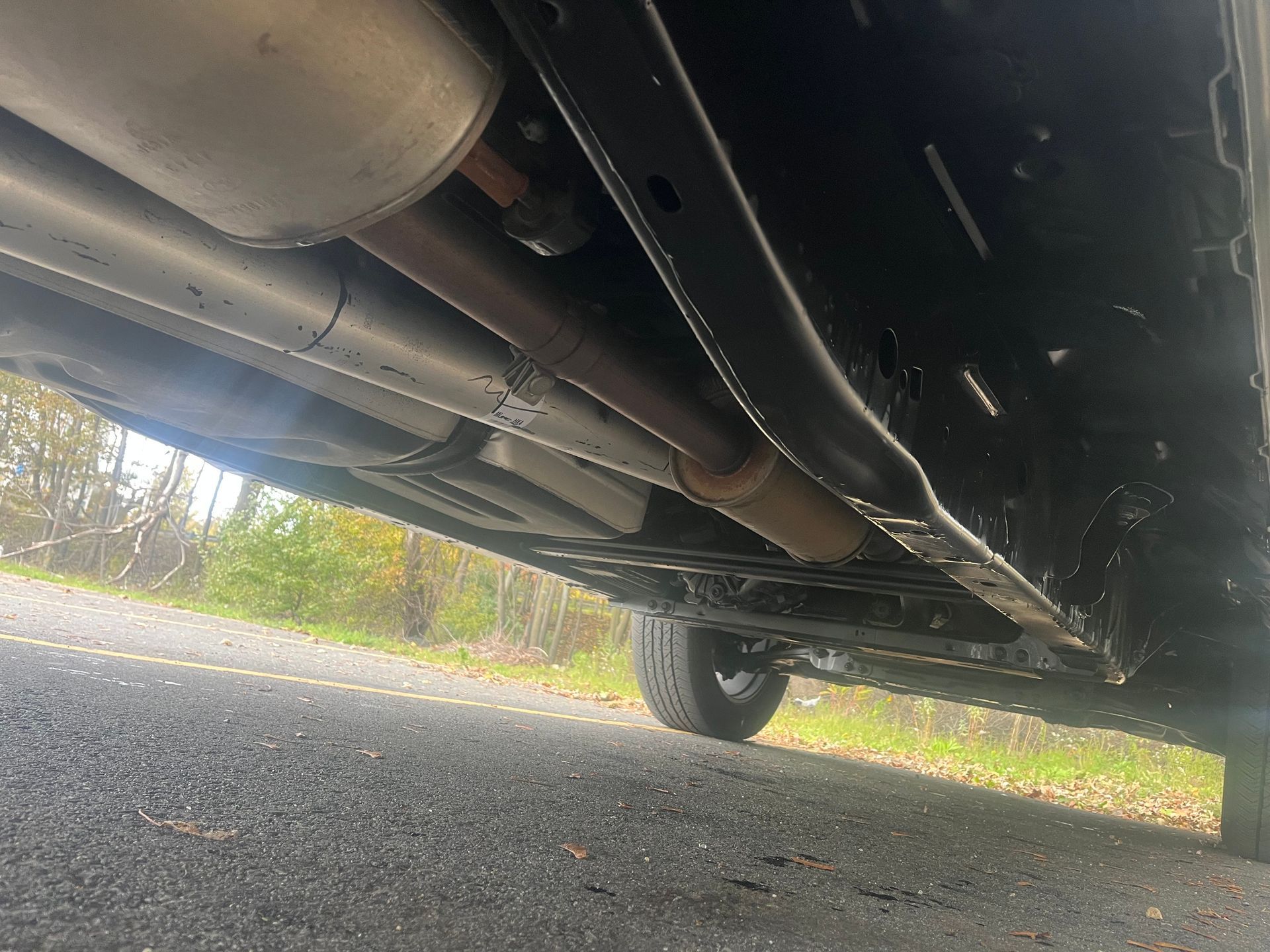 Undercarriage of a vehicle, showing exhaust system, drive shaft, frame, and tire. Black and silver components.