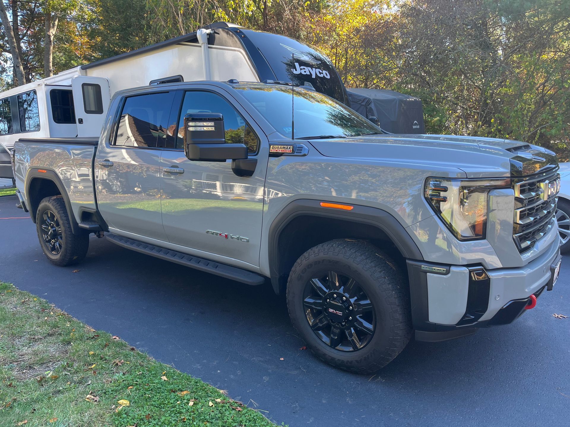 Gray GMC truck towing a white and black RV parked on a driveway in front of green grass and a tree.