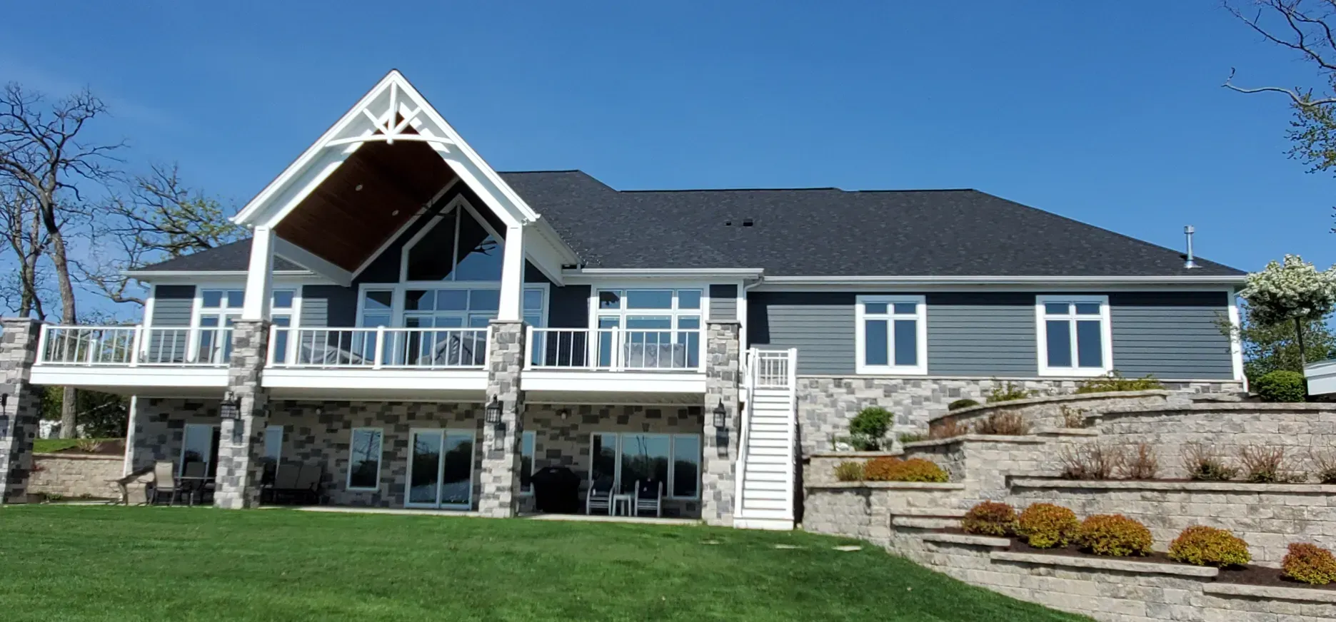 Large gray house with a wraparound porch, stone foundation, and landscaped lawn under a blue sky