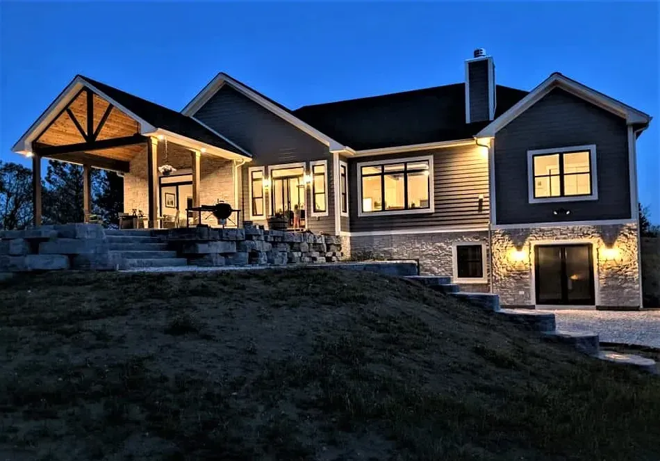 Illuminated stone house at dusk with lit windows and a covered porch on a sloped lawn