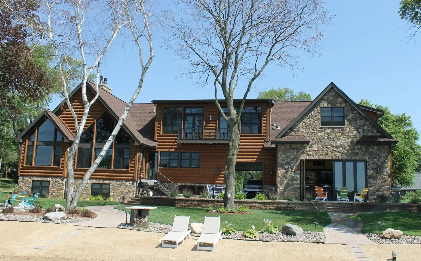 Rustic lakeside lodge with stone and wood facade, large windows, and two lounge chairs on sandy shore.