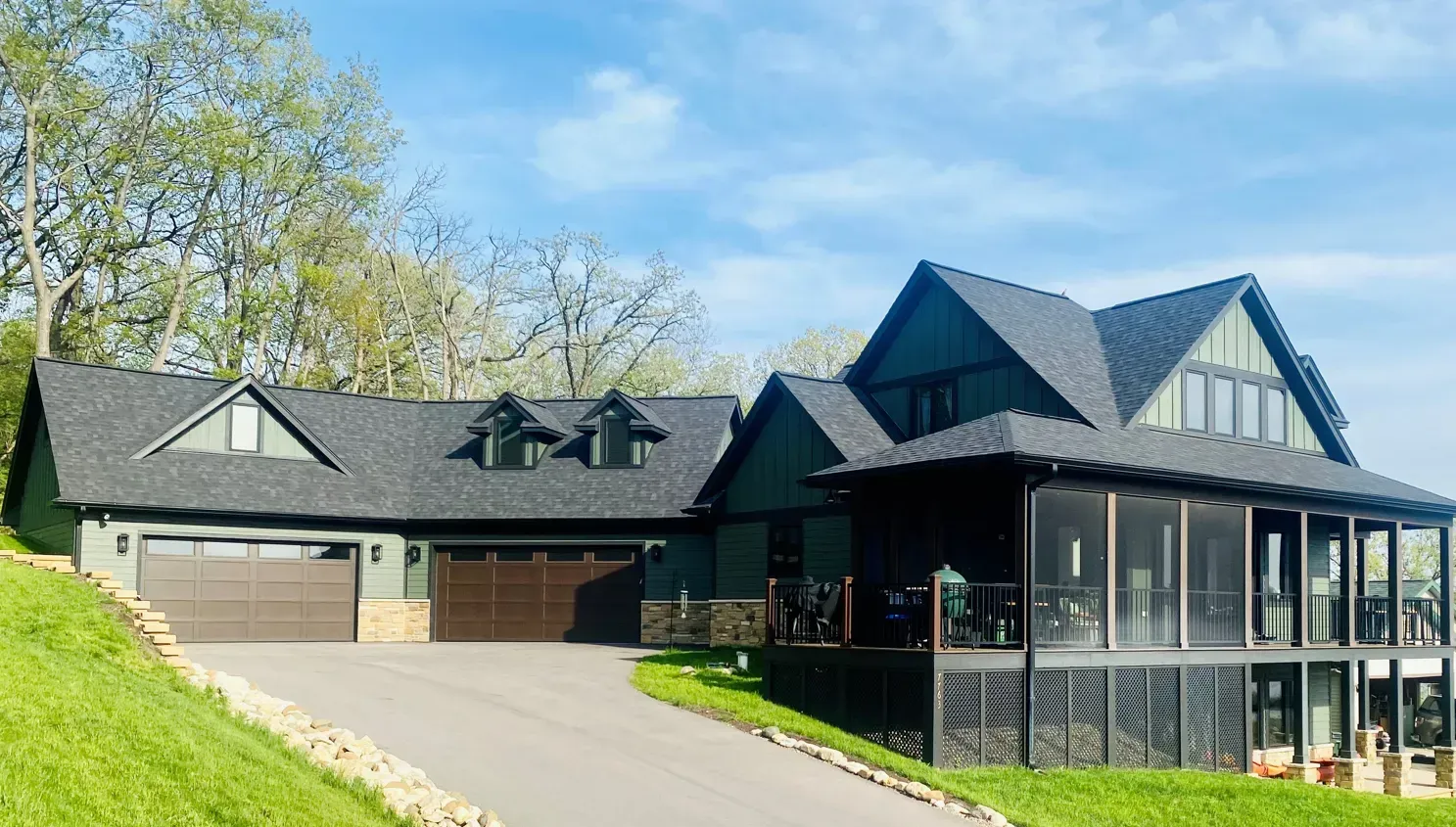 Modern black house with gabled roof, stone garage, and front porch on a sunny hillside.