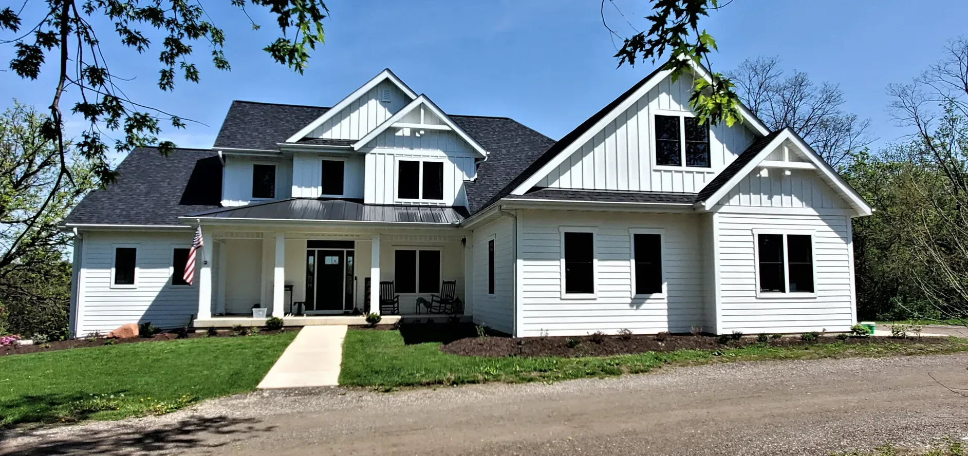 White farmhouse with black roof and porch, front walkway, and blue sky on a sunny day