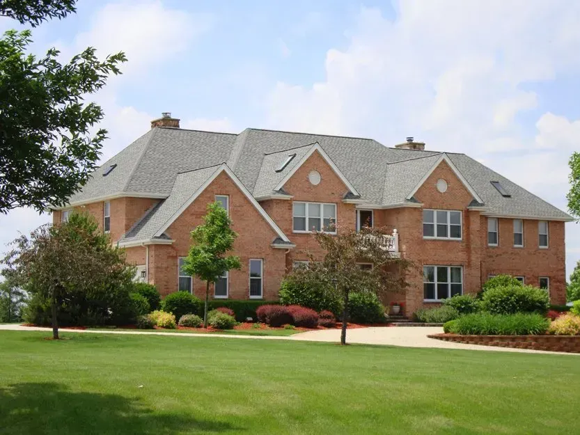 Large brick suburban house with gray roof, green lawn, and trees under a partly cloudy sky