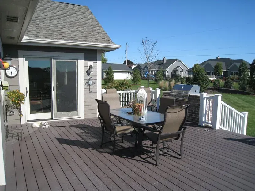 Backyard deck with patio table, chairs, and white railing beside a house on a sunny day