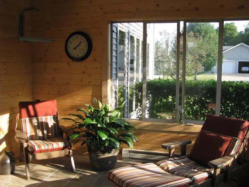 Sunlit wooden sunroom with two striped lounge chairs, a potted plant, and large windows overlooking a garden