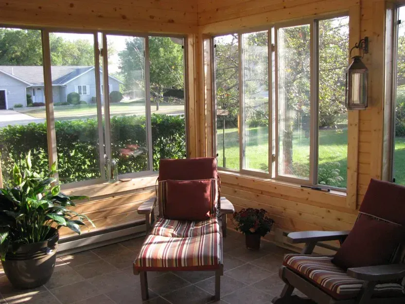 Sunroom with large windows, cushioned chairs, and potted plants overlooking a green yard