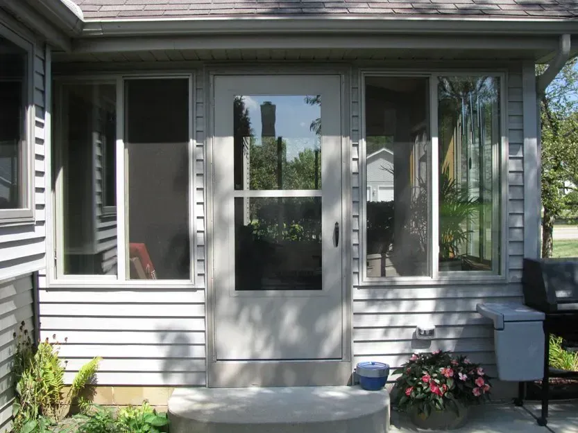 Front porch with white screen door, windows, a grill, and potted flowers on a sunny house exterior