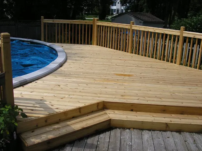 Wooden deck with railing beside a curved blue swimming pool outdoors