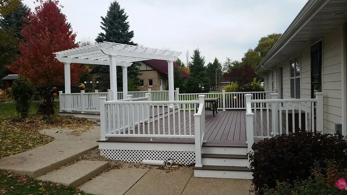 White wooden deck and pergola in a landscaped yard beside a house under overcast skies