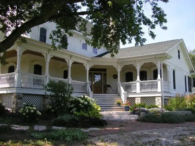White Victorian house with wraparound porch, stone steps, and a front garden shaded by trees
