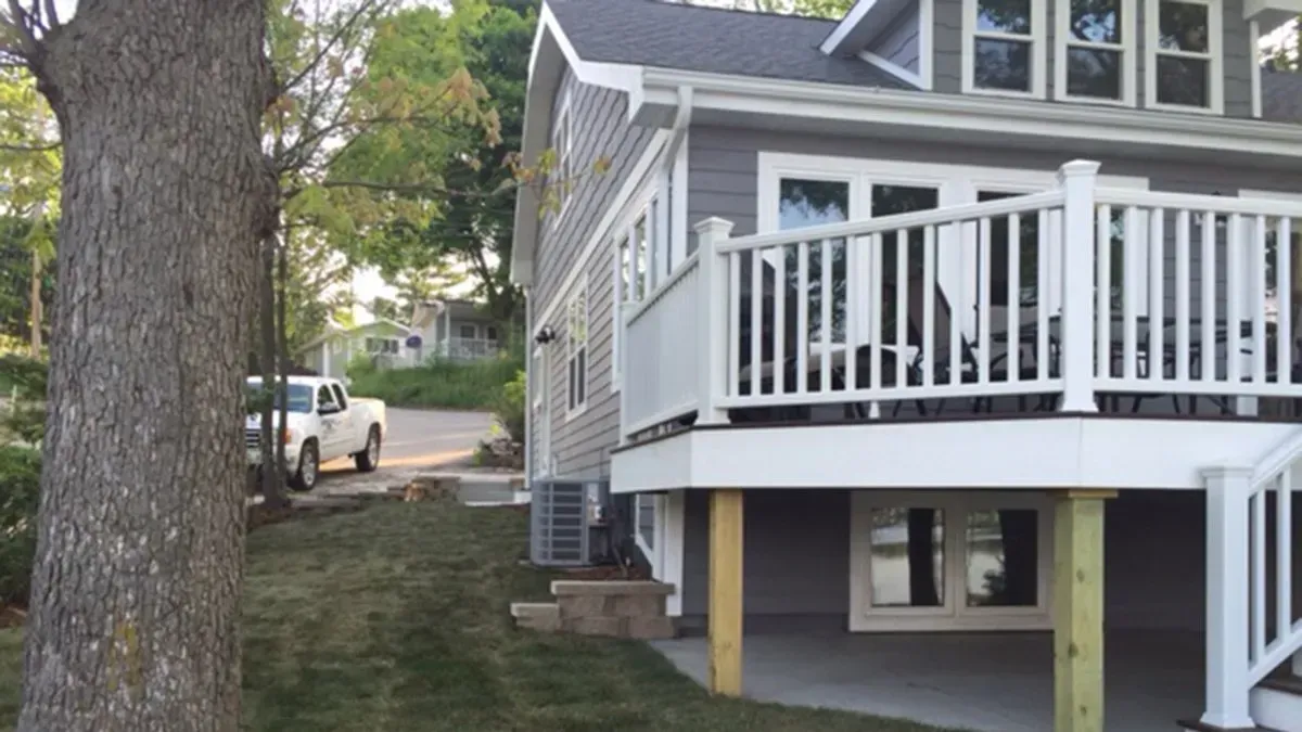 House with white deck beside a large tree and a driveway with a parked truck