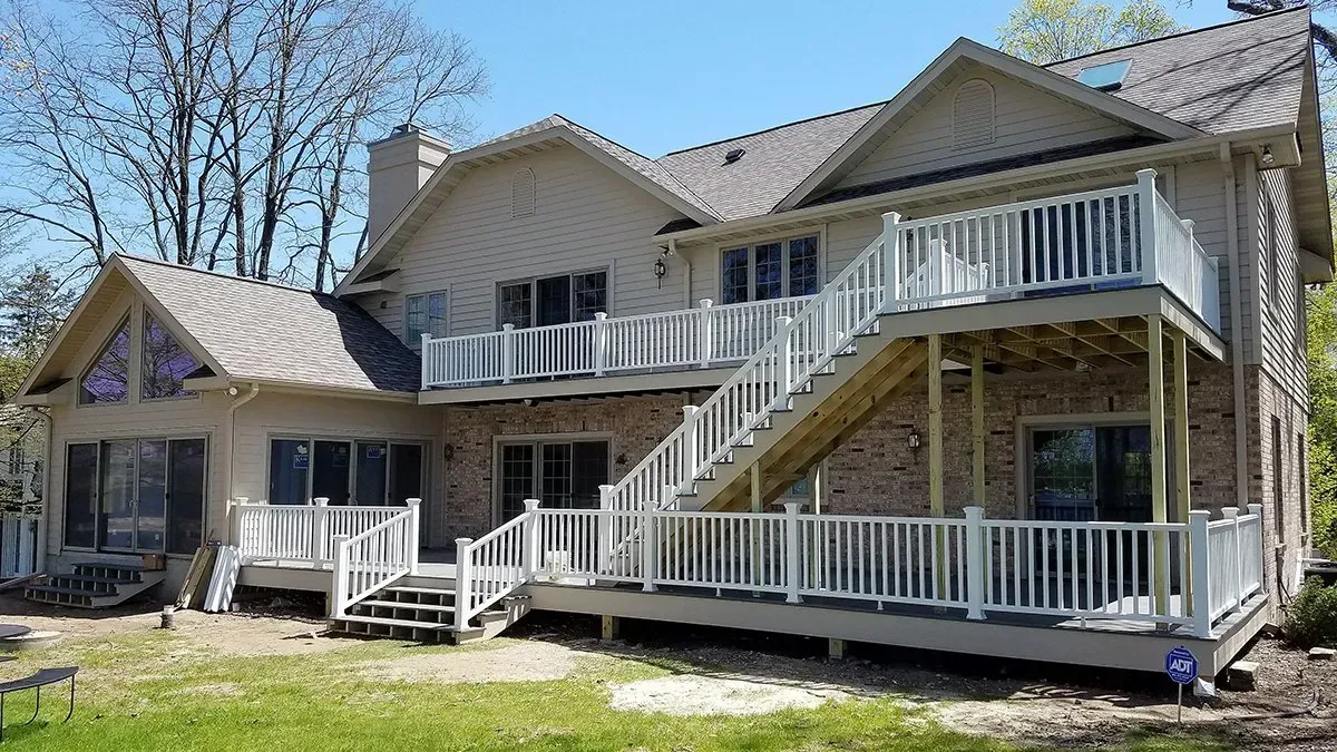 Two-story beige house with white railings and large wraparound decks in a grassy yard