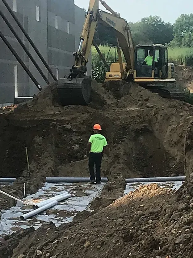 Worker in neon green vest stands by excavator digging a large dirt pile at a construction site.