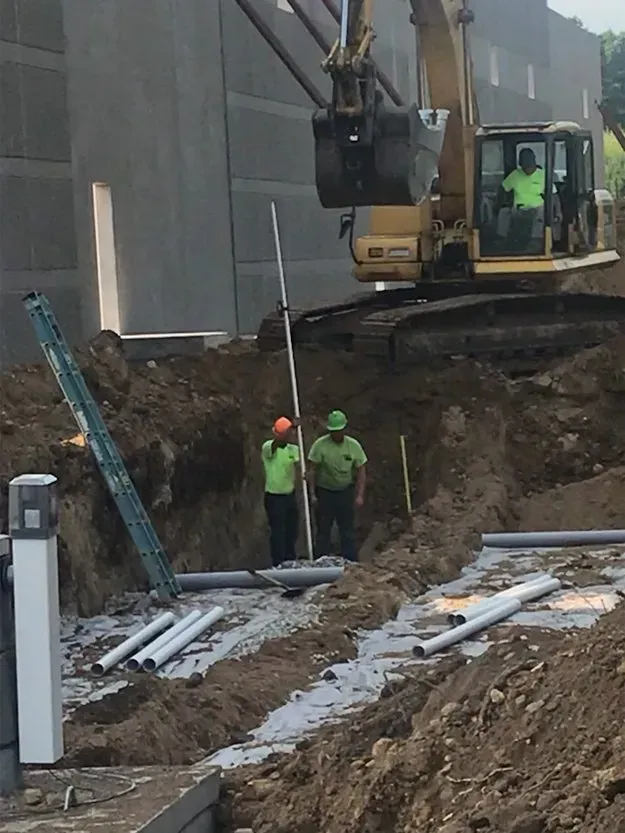 Construction site with excavator digging a trench beside a gray building; workers in safety vests nearby