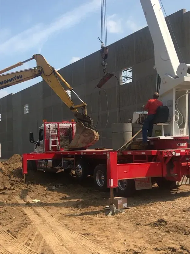 Excavator lifting debris into a red flatbed truck at a construction site beside a gray building