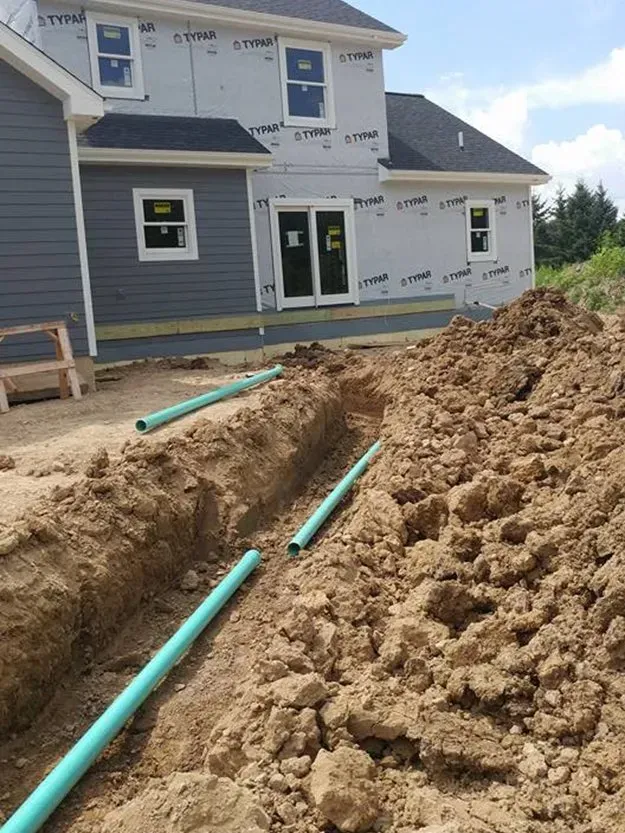 Backyard construction site beside a gray house, with dirt piles and green utility pipes in a trench