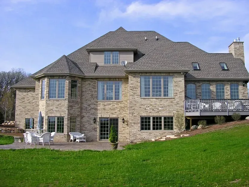 Large stone house with gray roof and patio on a green lawn under a blue sky