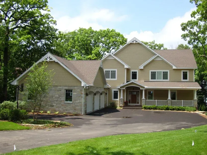 Large beige suburban house with stone garage, curved driveway, and green trees under a blue sky