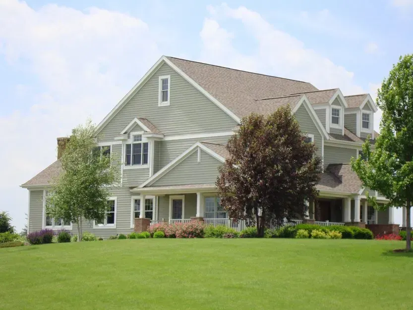 Large gray suburban house with a green lawn and trees under a blue sky