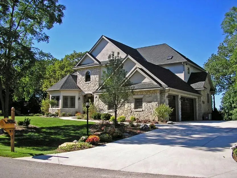 Large stone suburban house with a gabled roof, front lawn, and long driveway in bright sunlight