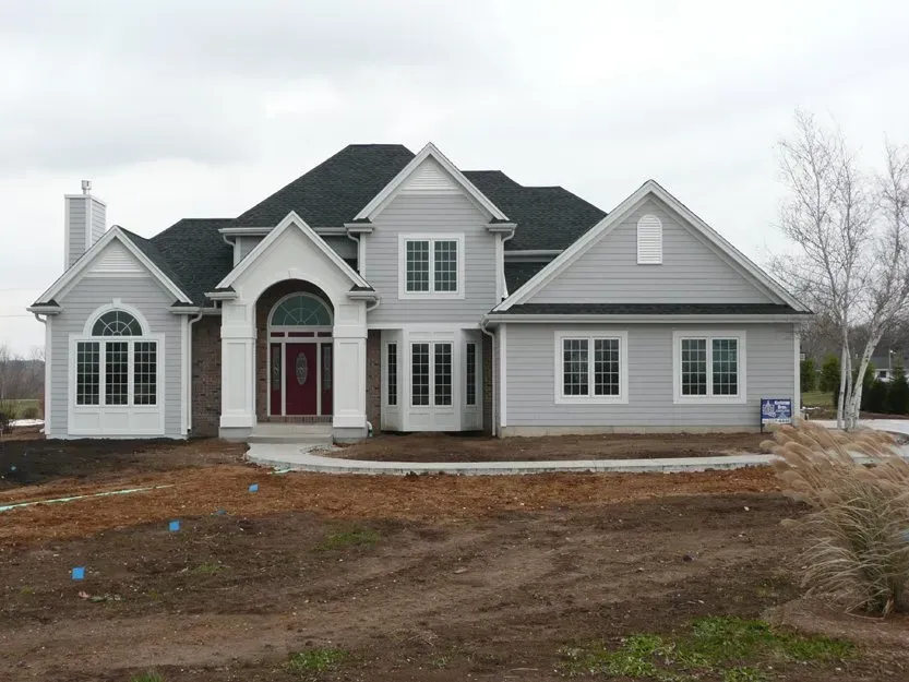 Large gray suburban house with dark roof and red front door, surrounded by bare dirt under an overcast sky
