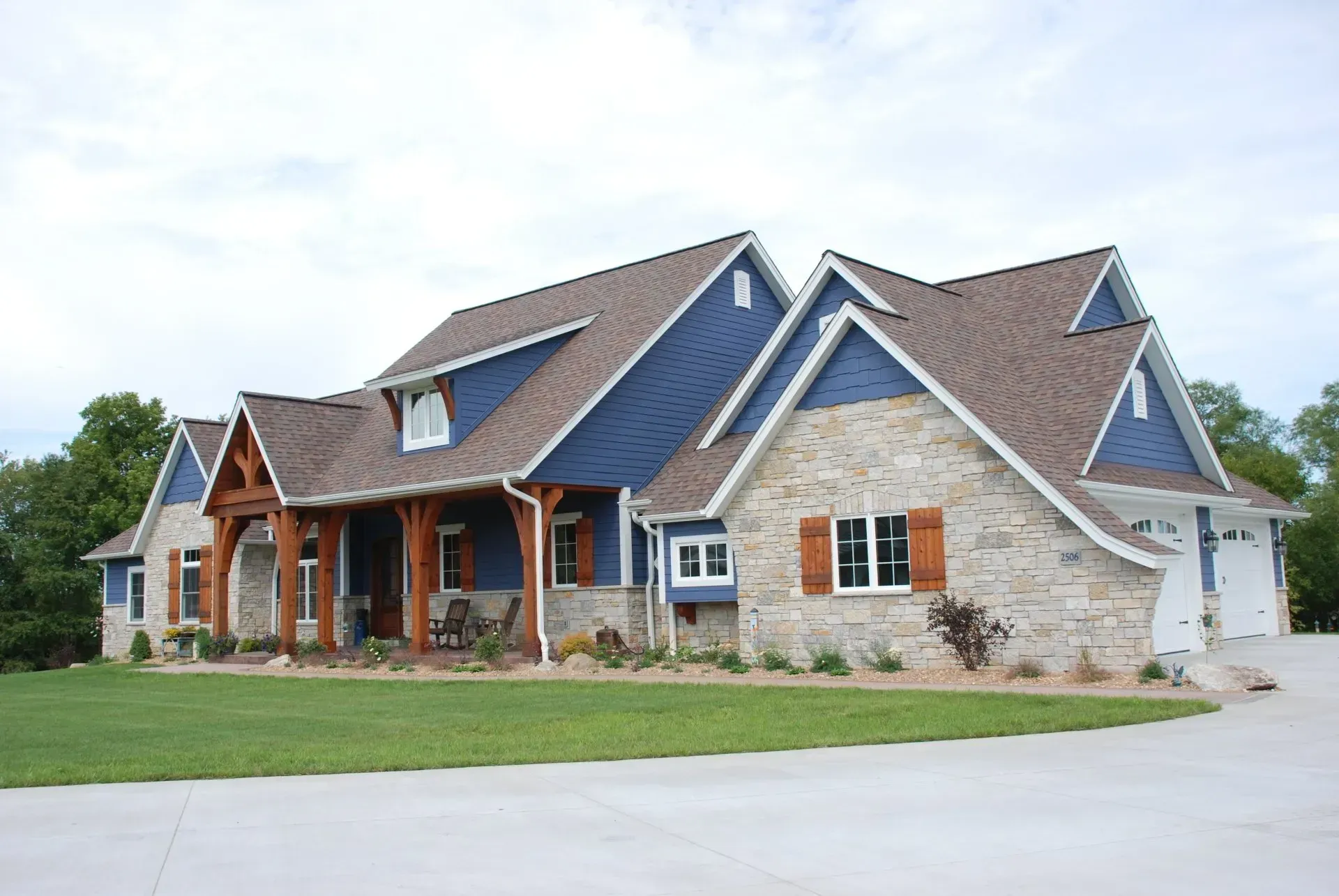 Large stone suburban house with a gabled roof, front porch, and attached garage on a green lawn.