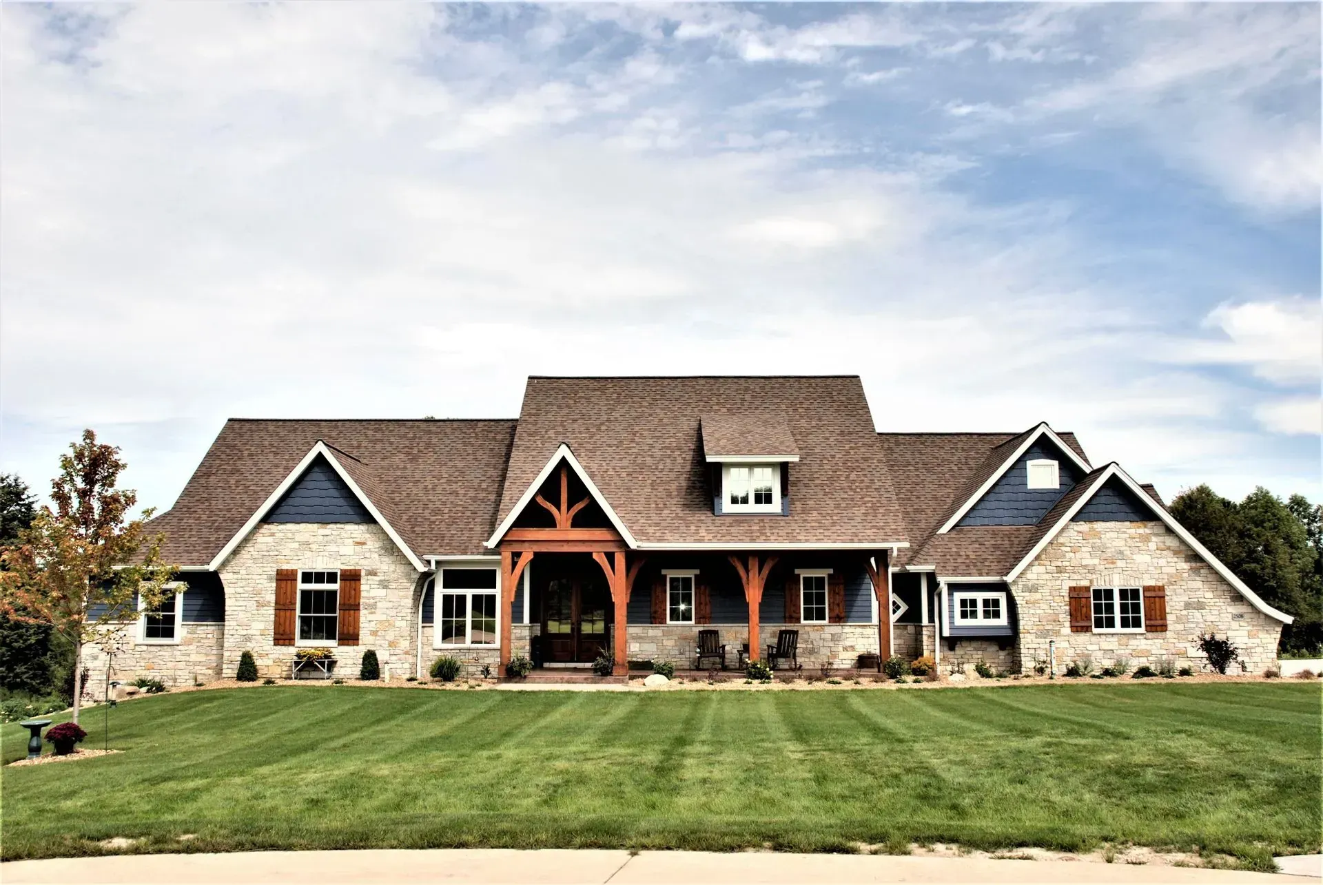 Large stone suburban house with multiple gables, a front porch, and a green lawn under a partly cloudy sky