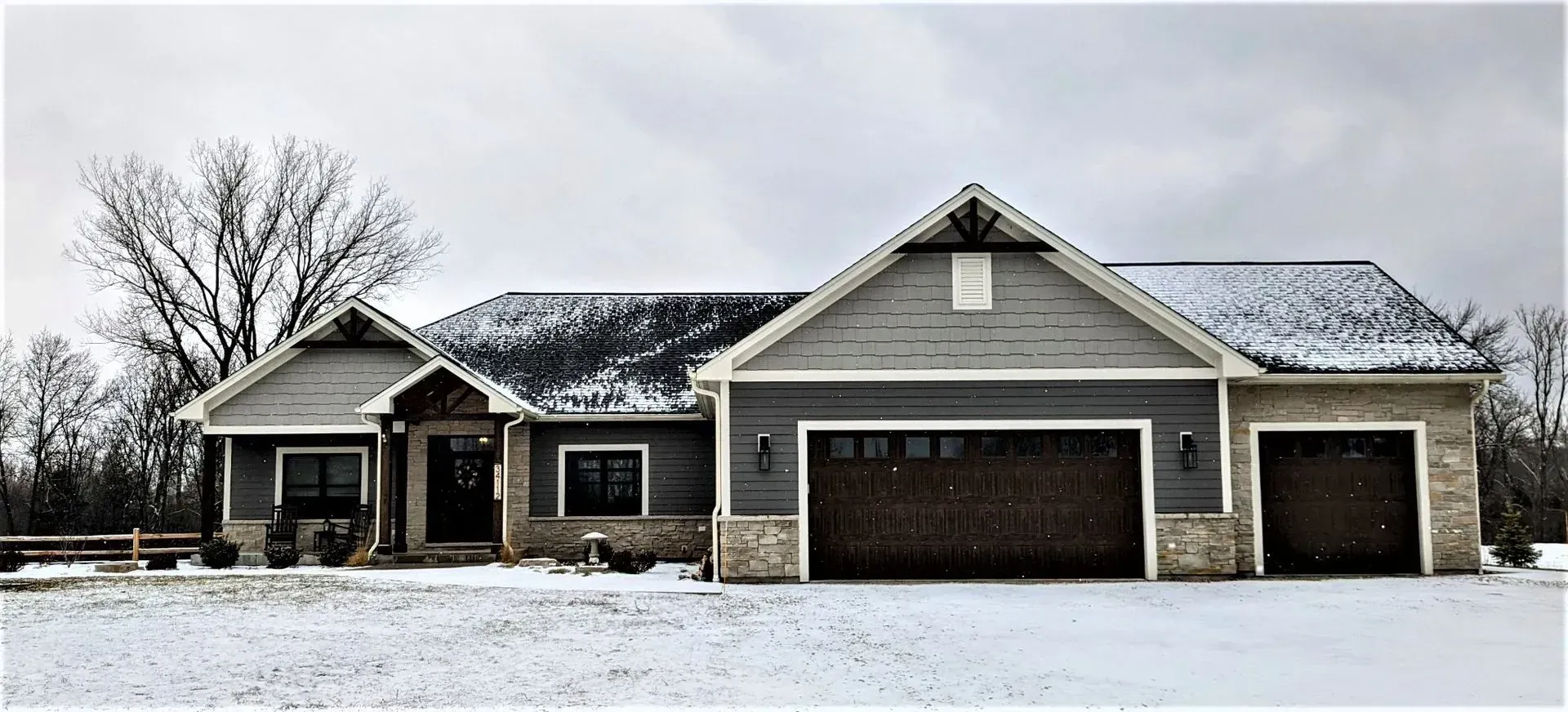 Snow-covered gray house with a three-car garage and bare tree in front