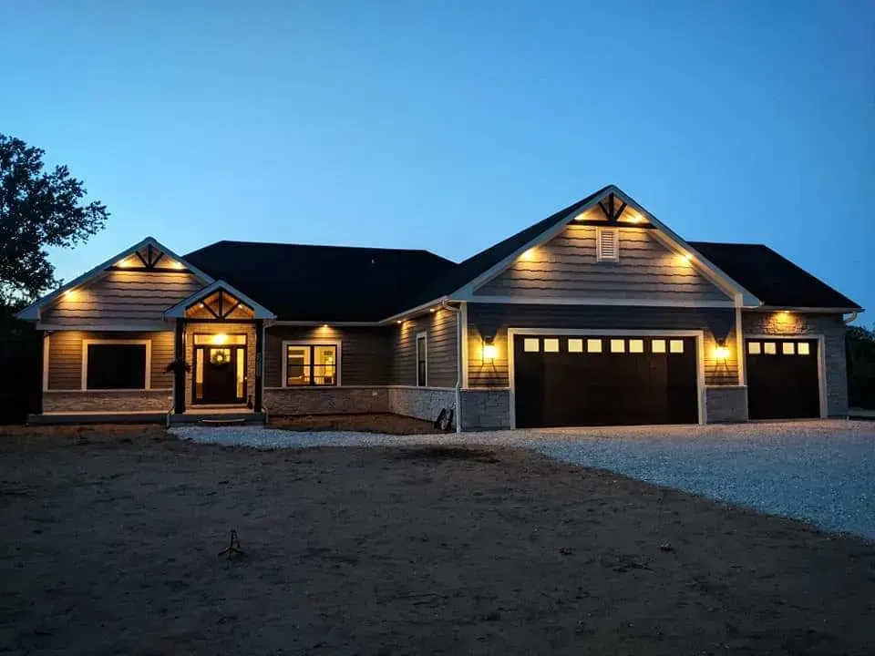 Single-story house at dusk with warm porch lights and a two-car garage on a dirt lot.