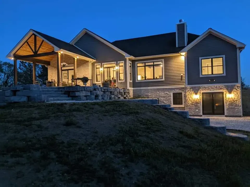 Large modern house at dusk with warm porch and window lights, set on a sloped yard.