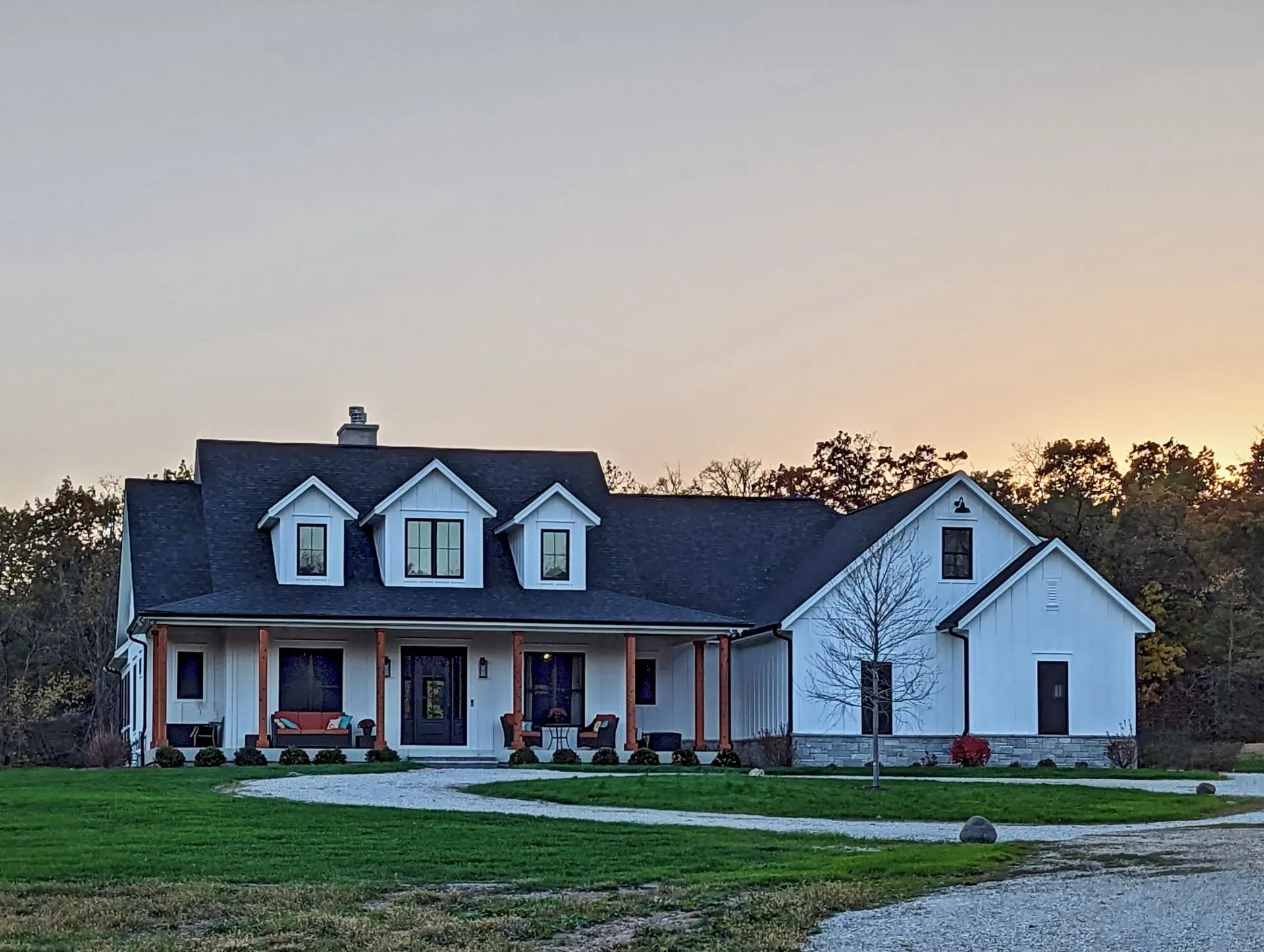 White farmhouse with front porch and dormer windows at dusk, set on a grassy lot under a pastel sky