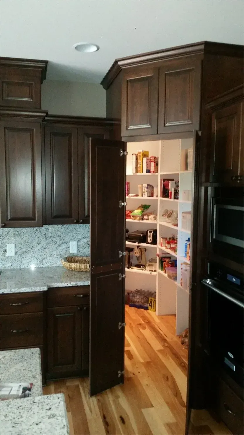 Kitchen doorway showing a lit pantry with shelves and hardwood floor between dark wood cabinets