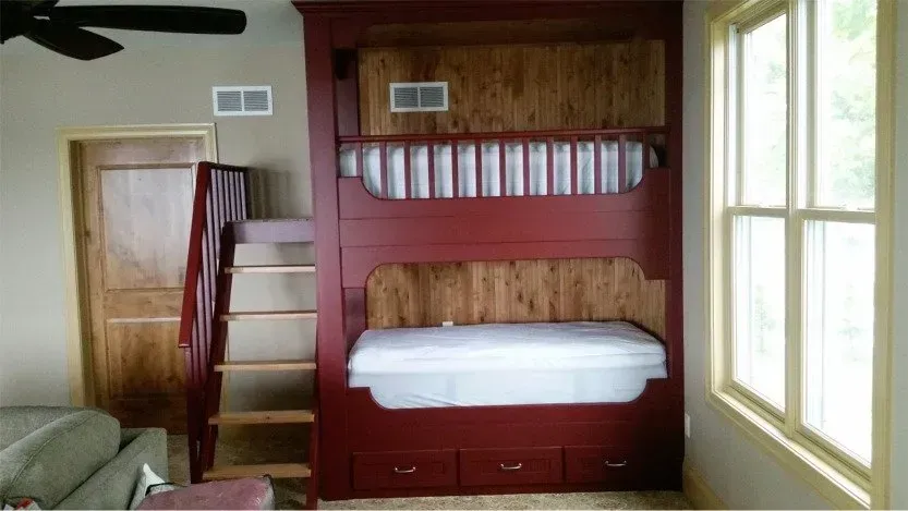 Bedroom with red bunk bed, white bedding, wooden doors, and a large window with sunlight.
