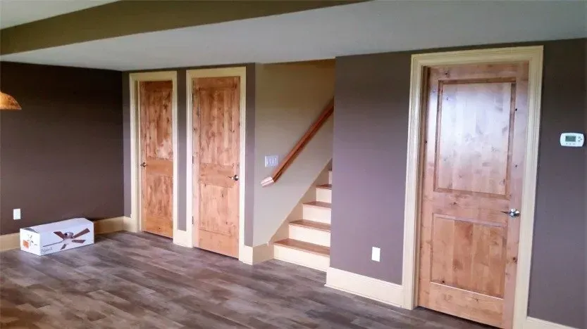 Basement room with wood floors, brown walls, three wood doors, and a small staircase.