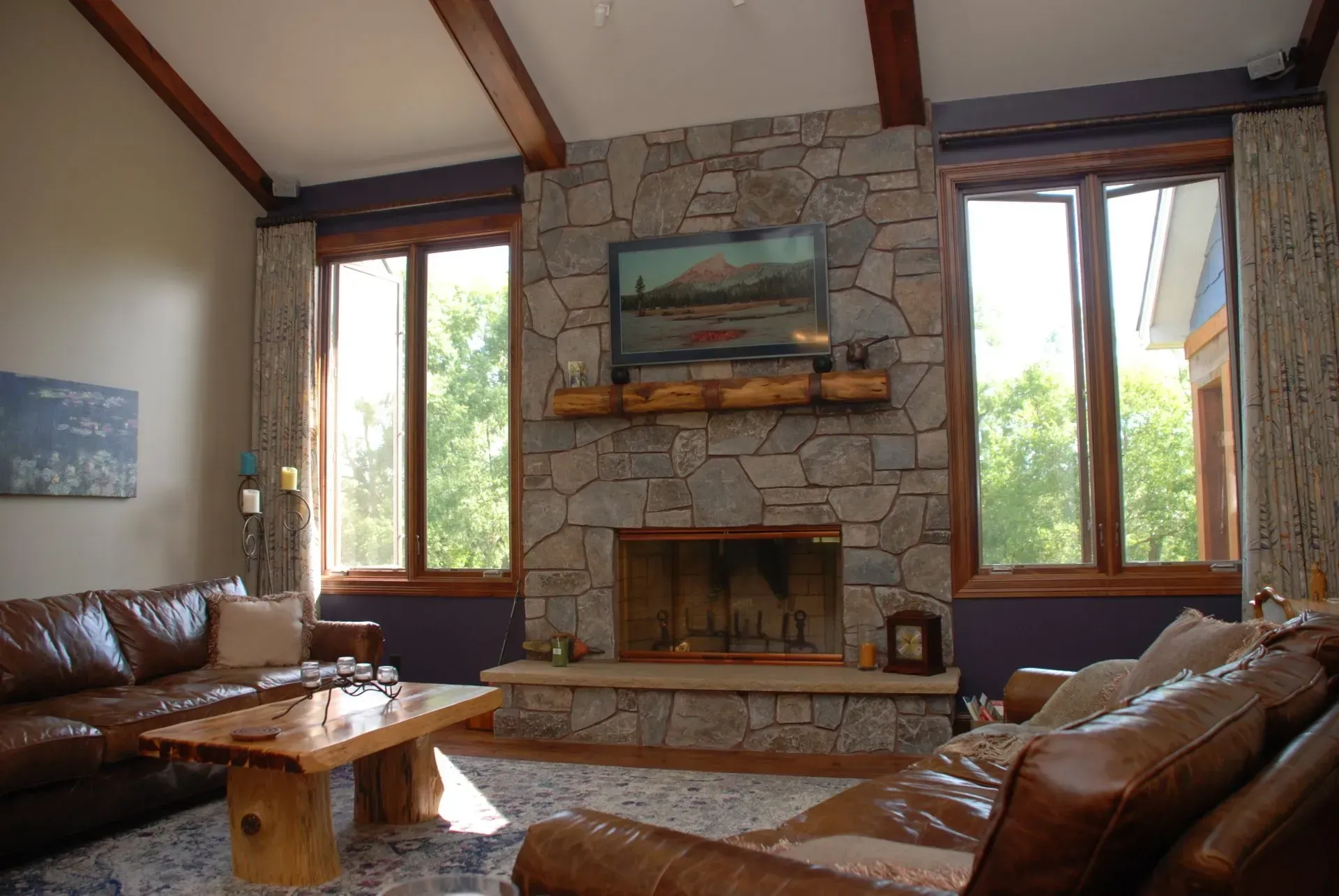 Rustic living room with stone fireplace, leather sofas, and large windows