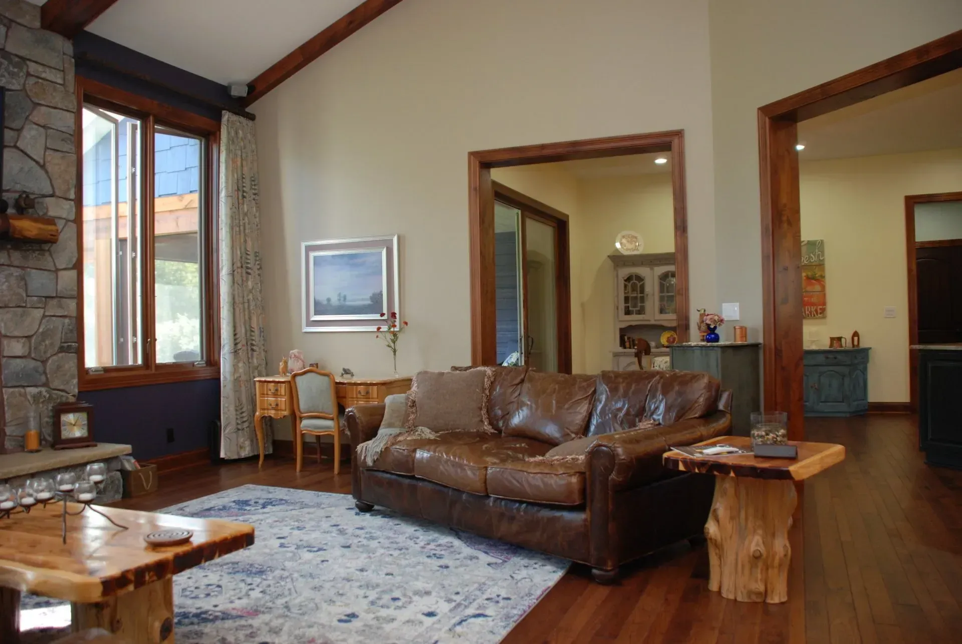 Spacious living room with a brown leather sofa, stone fireplace, wooden floors, and large windows.