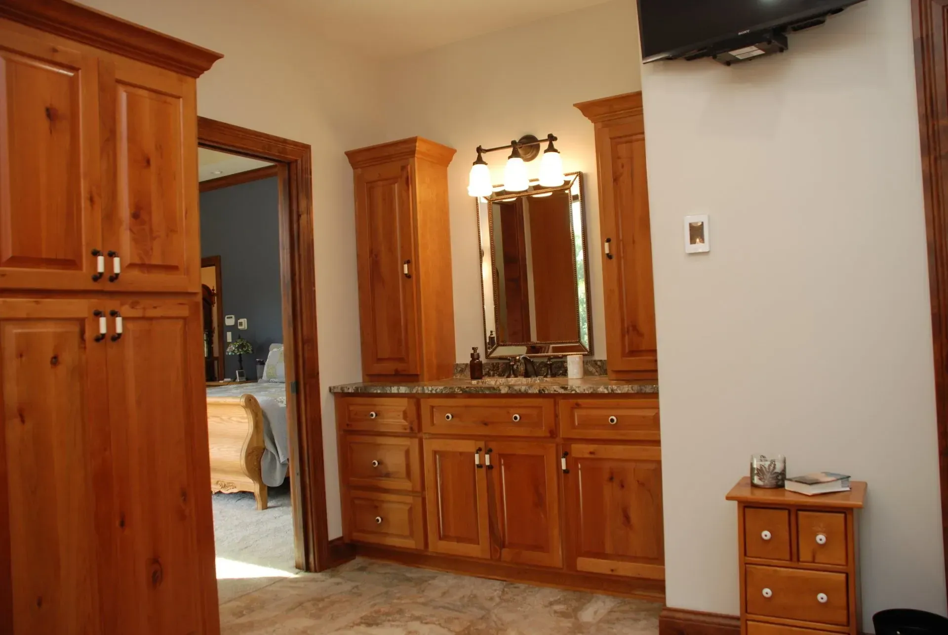 Wooden bathroom vanity with mirror and cabinets in a tiled room; a doorway opens to a bedroom.