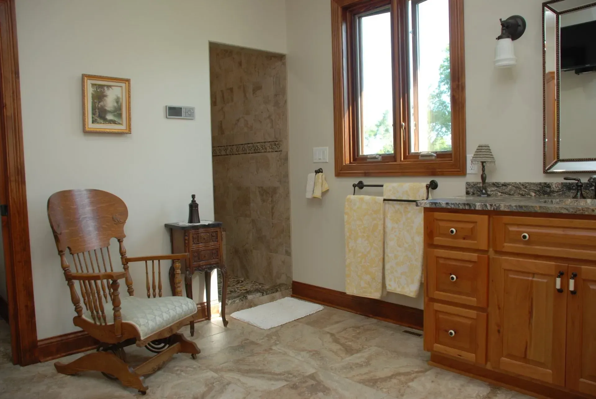 Bathroom interior with wood vanity, mirror, window, and a wooden chair by a tiled shower area