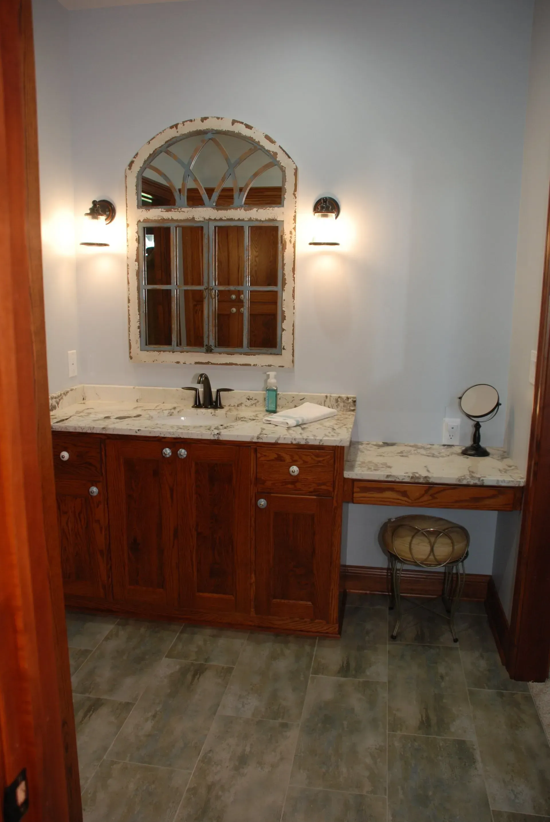 Bathroom vanity with marble countertop, arched mirror, wall sconces, and a small stool beneath the sink.