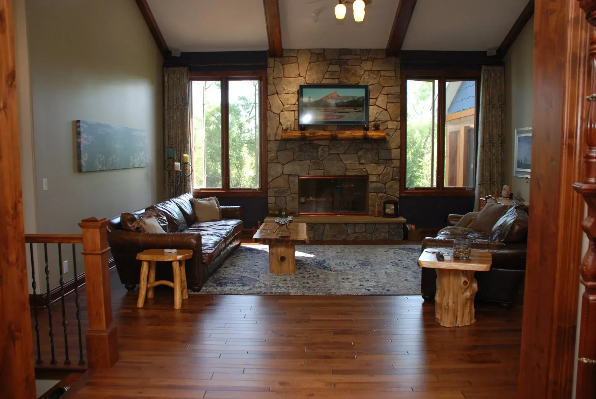 Rustic living room with stone fireplace, leather sofas, wood floors, and large windows