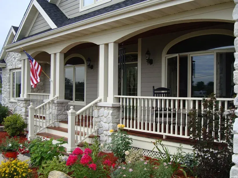 Front porch of a house with white railings, an American flag, and red flowers in the garden.