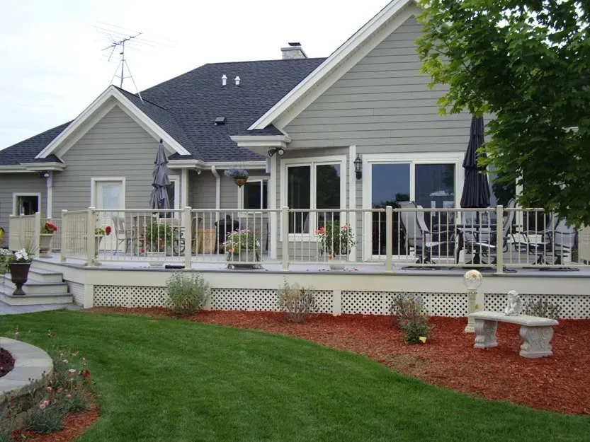 Gray house with white deck railing, red mulch landscaping, and green lawn