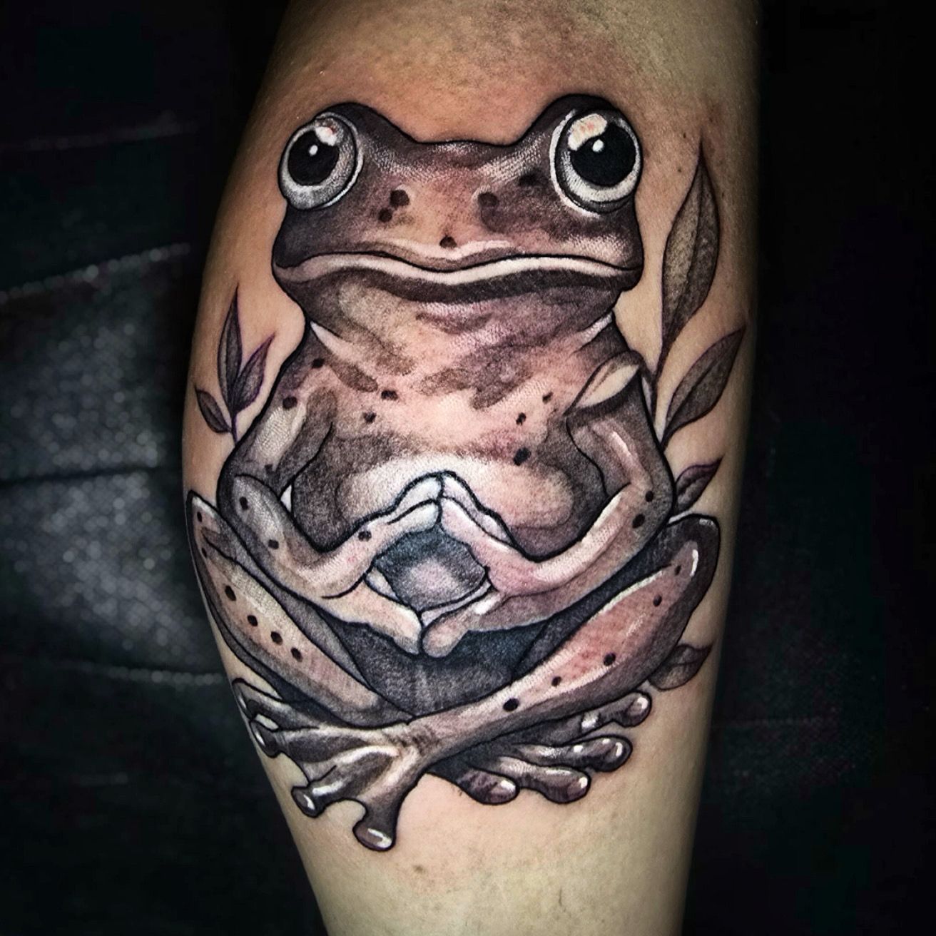 Tattoo of a frog with large eyes, arms crossed, and legs folded. Black and grey ink on skin.