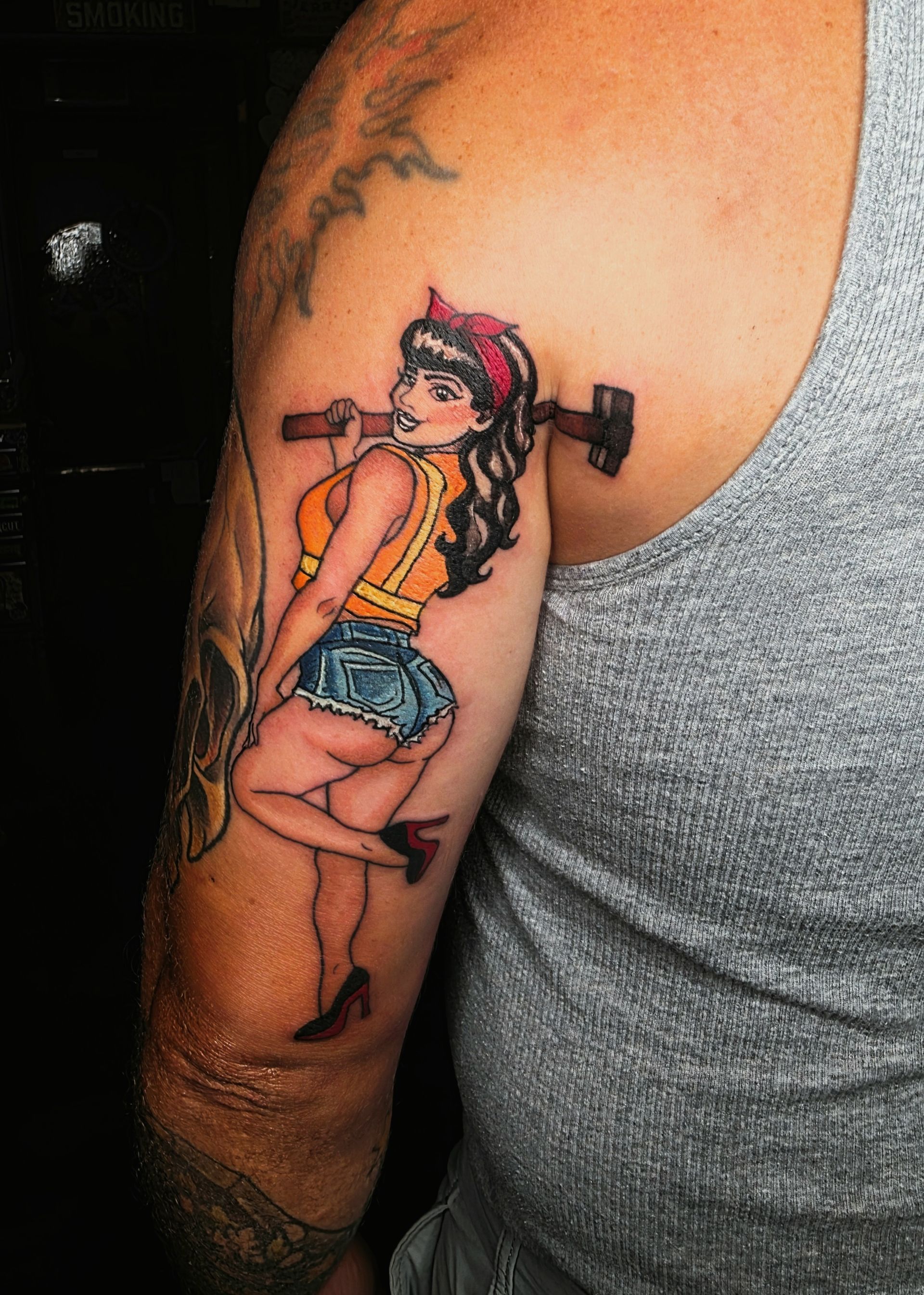 Tattoo on arm of a woman holding a hammer, wearing work clothes, a red headband, and high heels.