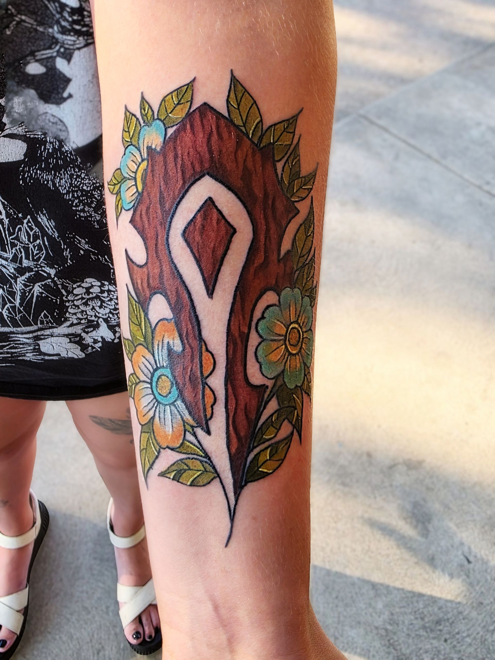 Arm tattoo featuring the Horde symbol from World of Warcraft, surrounded by flowers and leaves; colorful design.