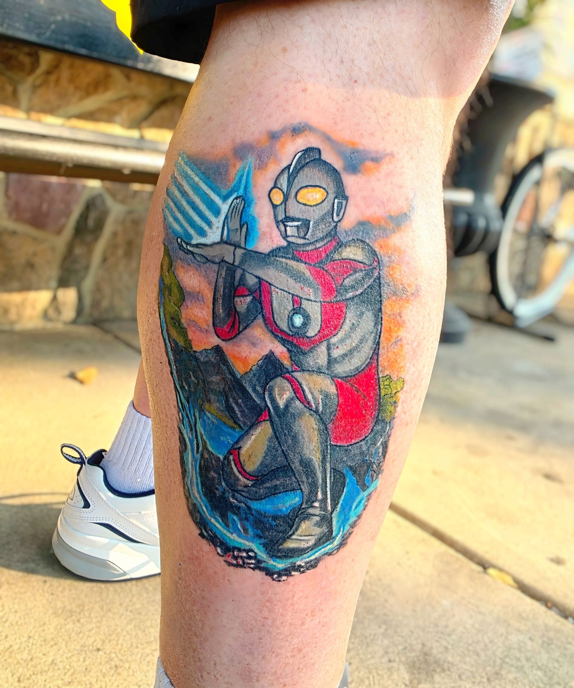 Ultraman tattoo on calf, posing with outstretched arms. Colorful backdrop of blue, orange, and green.