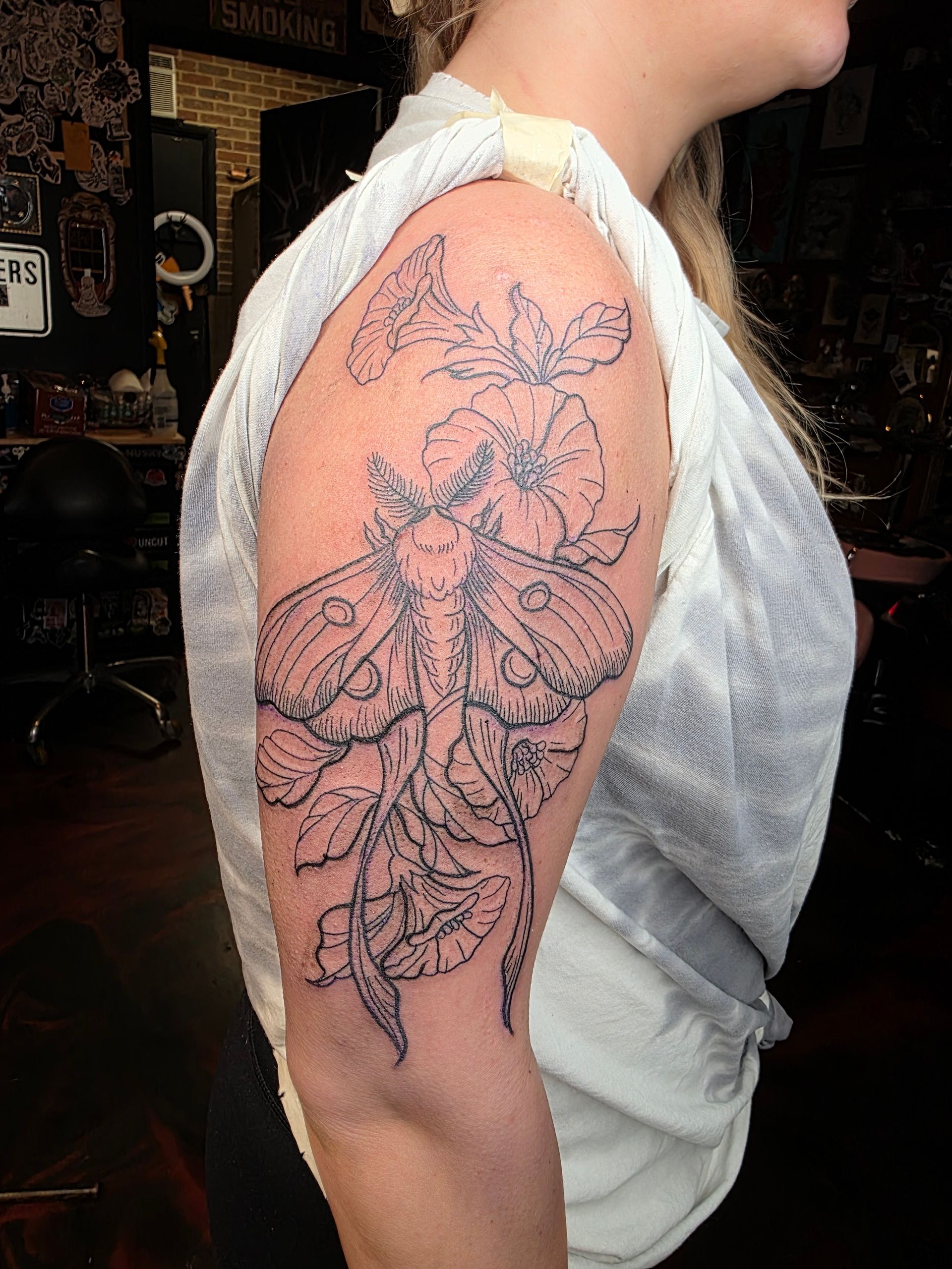 Tattoo of a moth with floral details on a person's upper arm. Black outline, indoor setting.