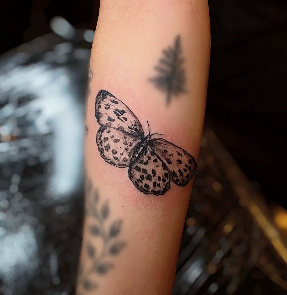 Black dotted butterfly tattoo on arm, with a blurry tree and leafy design.